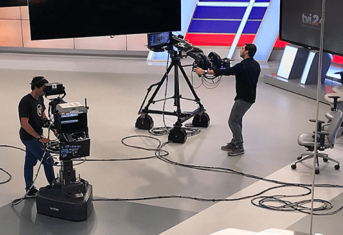 Two cameramen in a TV studio operate professional cameras on stands, with large screens and color panels in the background.
