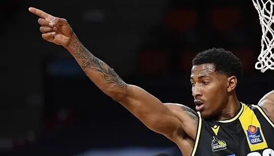 Basketballspieler in schwarzem Trikot mit gelben Streifen hebt den Arm und zeigt in eine Richtung. Sichtbar sind tätowierter Arm und Spielfeld im Hintergrund.