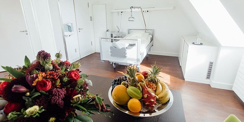 Modernes Patientenzimmer mit weißem Bett und medizinischer Ausstattung. Im Vordergrund ein Tisch mit einem Blumenstrauß und einer Schale voller frischer Früchte wie Ananas, Bananen und Äpfel.