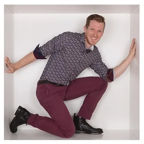 Andreas Klose Mitarbeiter von RIEDEL Networks befindet sich in einer weißen Box und kniet mit einem Bein auf dem Boden, während das andere angewinkelt ist. Die Arme sind seitlich ausgestreckt und stützen sich an den Wänden. Andreas trägt ein gemustertes Hemd.